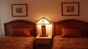 Una habitación con dos camas, cada una con cabecero de madera, una mesita de noche entre ellas y una lámpara de mesa. Hay cuadros enmarcados en las paredes.