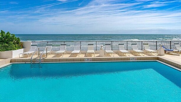 Una piscina rectangular con agua cristalina azul, rodeada por una terraza blanca con sillas de descanso y una barandilla metálica.