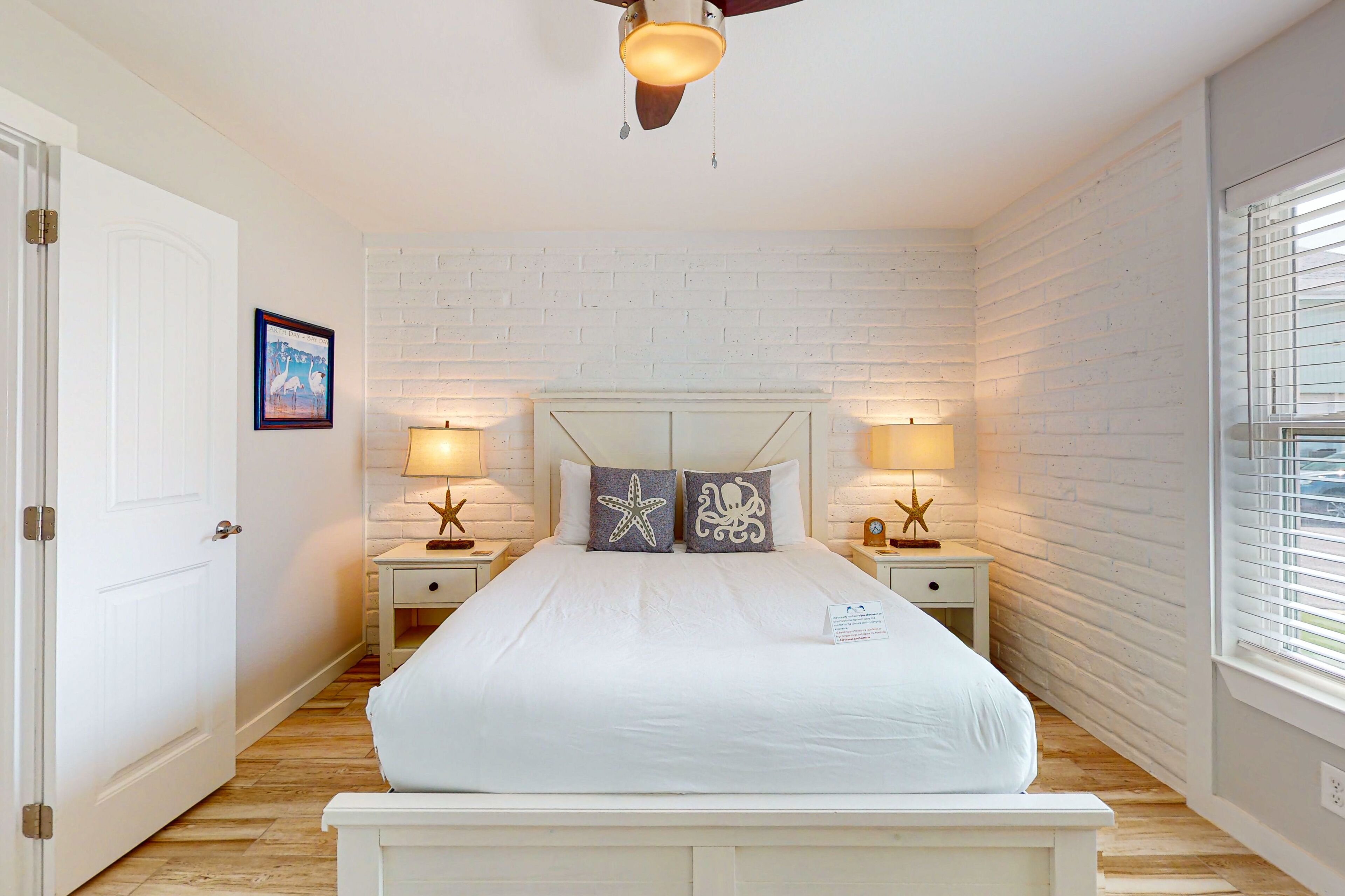 A bedroom with a white brick wall, a wooden floor, a white bed with two decorative pillows, two bedside tables with lamps, and a ceiling fan.