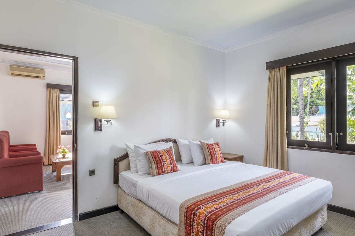 A hotel room with a large bed, two bedside tables with lamps, a red armchair, and a view of greenery through the window.