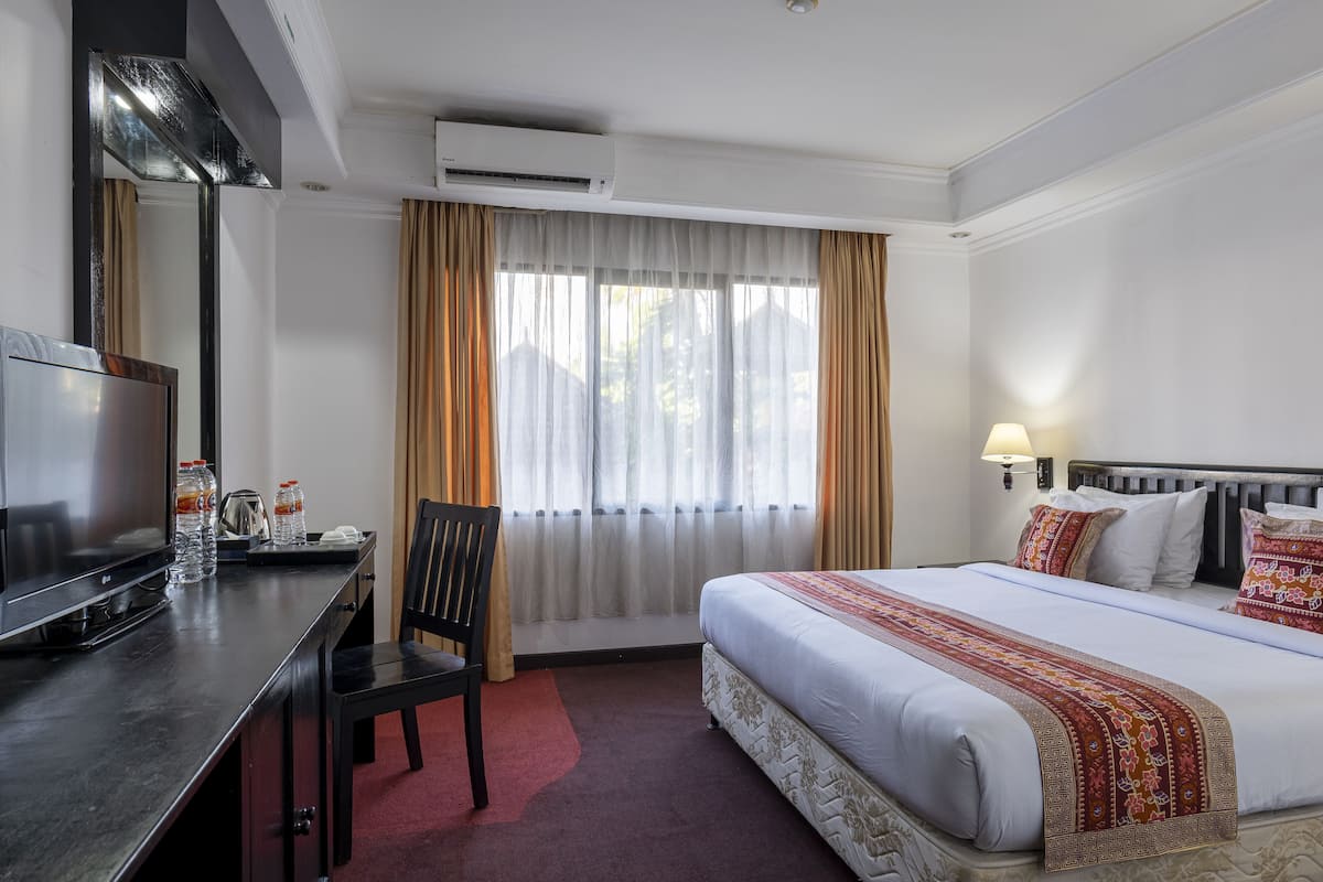 A hotel room with a bed, a desk with a TV, a chair, a window with curtains, and an air conditioning unit.