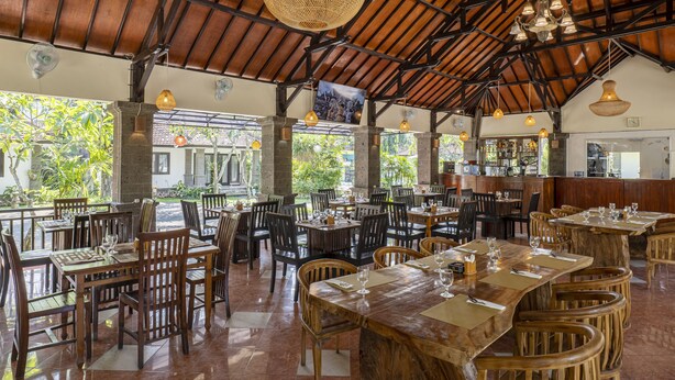A spacious restaurant with wooden tables and chairs, a high ceiling with exposed beams, and large windows offering views of greenery outside. - Jimbarwana Hotel (Negara)