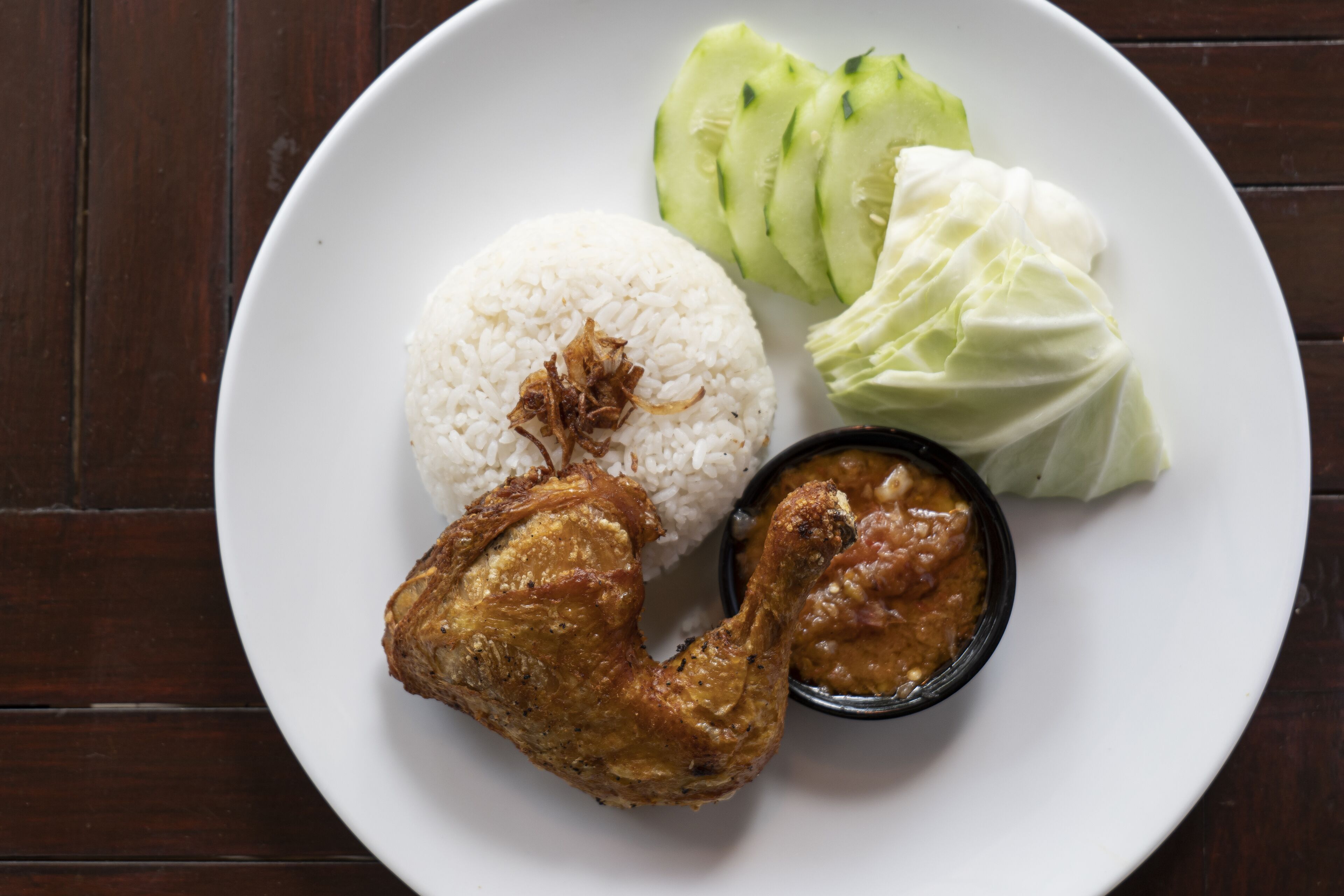 A plate with white rice, fried chicken, cucumber slices, cabbage, and a small bowl of sauce.