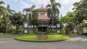 A hotel with a central entrance, surrounded by a landscaped garden with palm trees and a paved driveway. - Jimbarwana Hotel (Negara)