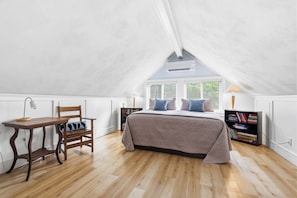 Un dormitorio con techo inclinado, una cama con ropa de cama estampada, una silla de madera, un escritorio con lámpara y una estantería.