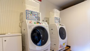 Deux machines à laver à chargement frontal, dotées de panneaux de commande, sont posées sur un plancher en bois dans une pièce dont les murs sont recouverts d’un papier peint à rayures verticales.