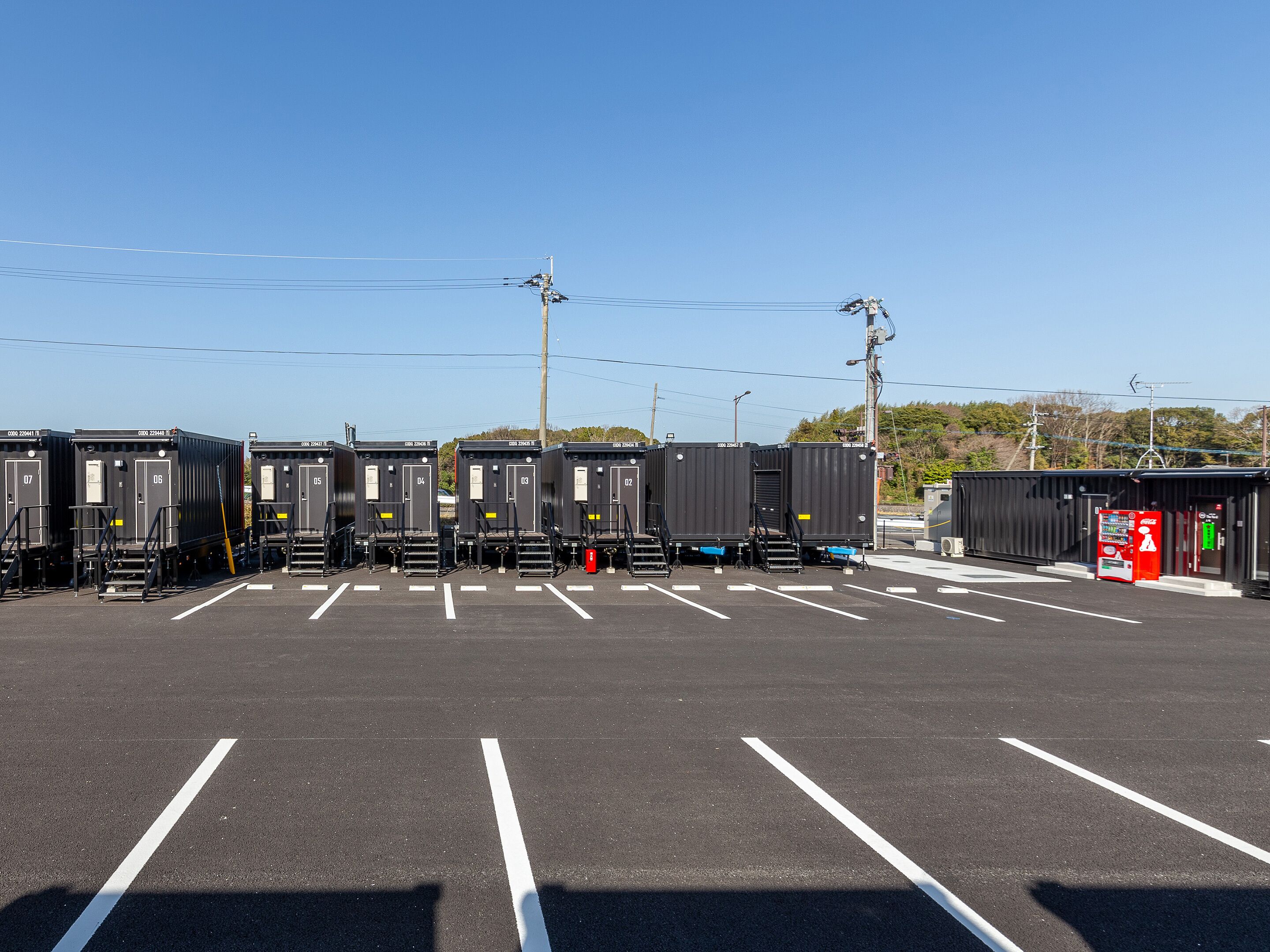 A parking lot with several black shipping containers, some with visible numbers and letters, and a red vending machine.