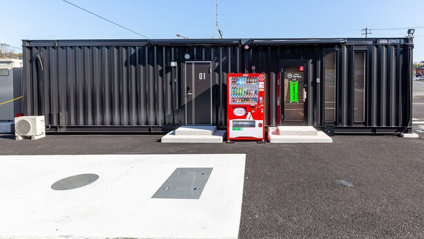 Un bâtiment en métal noir avec un distributeur automatique de Coca-Cola, une porte avec une pancarte et un couvercle de bouche d’égout sur le sol.