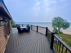 A wooden deck with a table and chairs, overlooking a beach and the ocean. - Lake Michigan Cabin + Hot Tub & Spectacular Views • Sleeps 8 (Manistique)