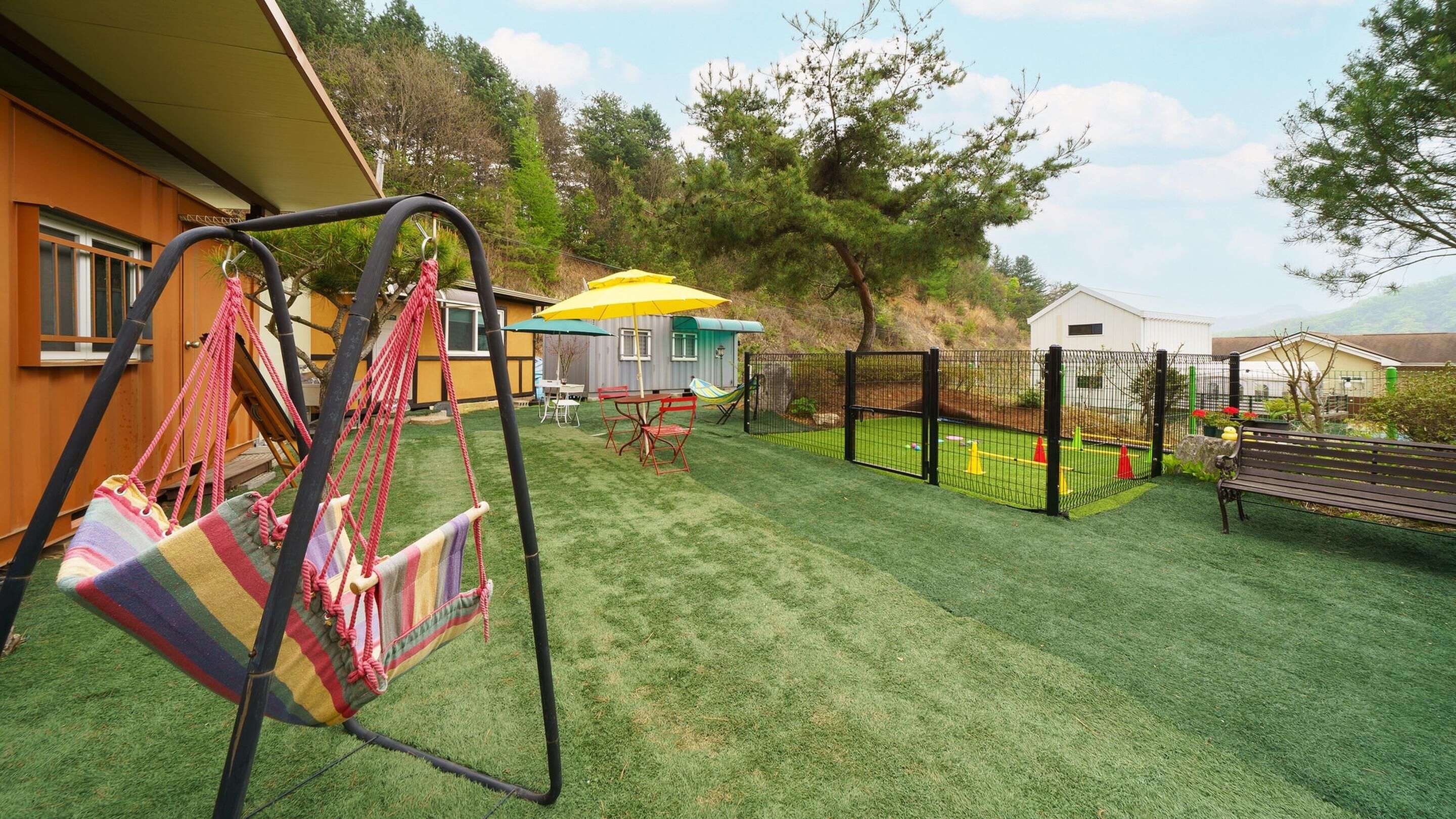 A backyard with artificial grass, a swing set, a play area with a yellow umbrella, and a fenced play zone.