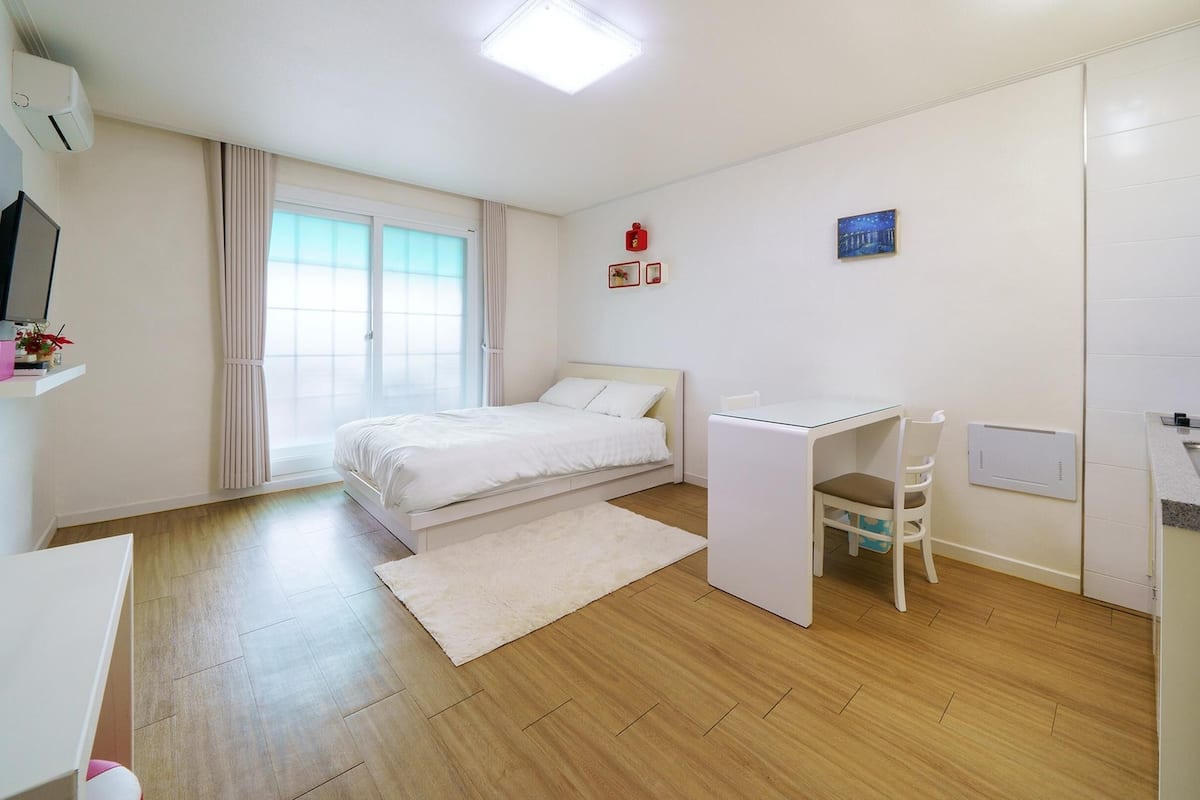 A bedroom with a bed, a desk, and a chair. There is a window with curtains, a TV on the wall, and a small shelf with items.