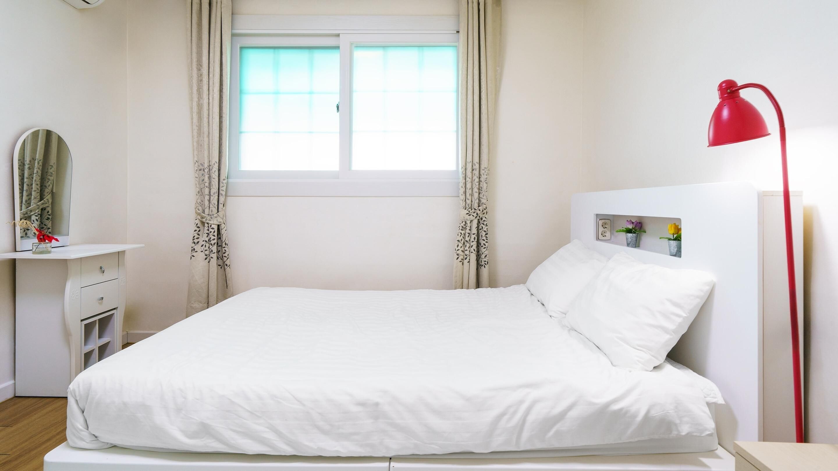 A neatly made bed with white bedding, a red floor lamp, a bedside table with a mirror, and a window with curtains.