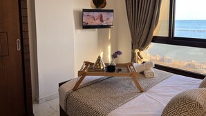 Een hotelkamer met een bed, een aan de muur bevestigde televisie, een klein tafeltje met een dienblad en uitzicht op zee.