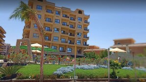 Een hotel met meerdere verdiepingen, balkons, een tuin met planten en parasols, en een heldere lucht.