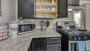 Cocina compacta con microondas, cafetera, estufa y alacena abierta que muestra vajilla y utensilios.