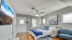 Un dormitorio con cama, una silla, ventilador de techo, ventana y un cuadro en la pared.