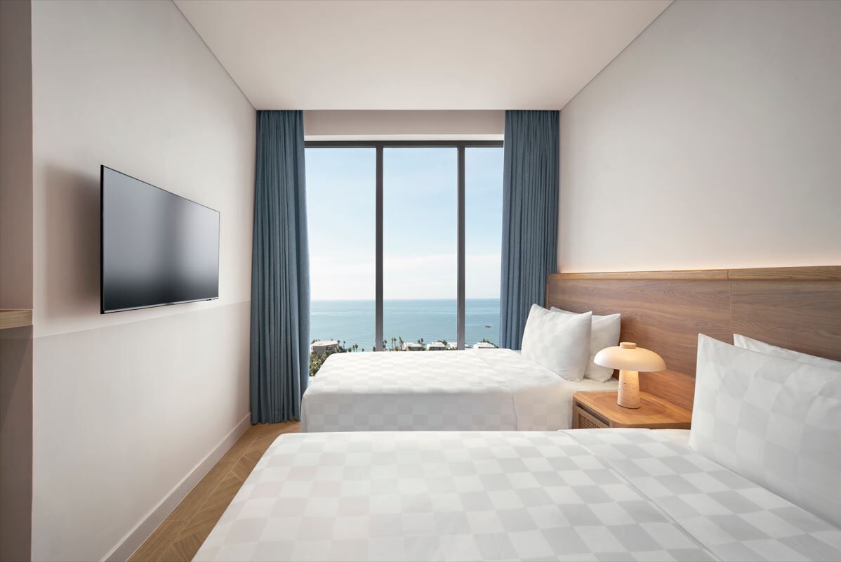 Habitació d'hotel amb un llit gran, una taula de nit amb una làmpada, un televisor de pantalla plana muntat a la paret i vistes a l'oceà a través de grans finestres.