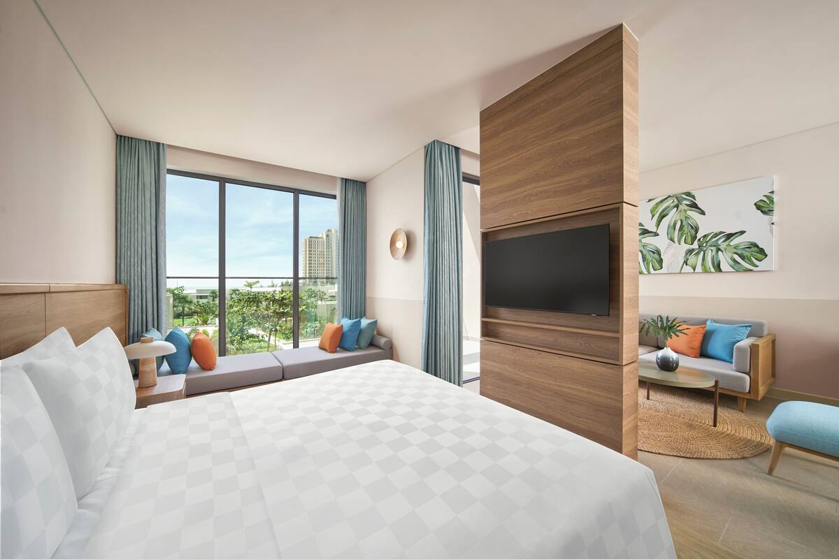 A modern hotel room with a large bed, a TV, a sofa, and a window with a city view.