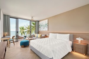A modern hotel room with a large bed, a seating area with a view of the ocean, a dining table, and a small balcony.