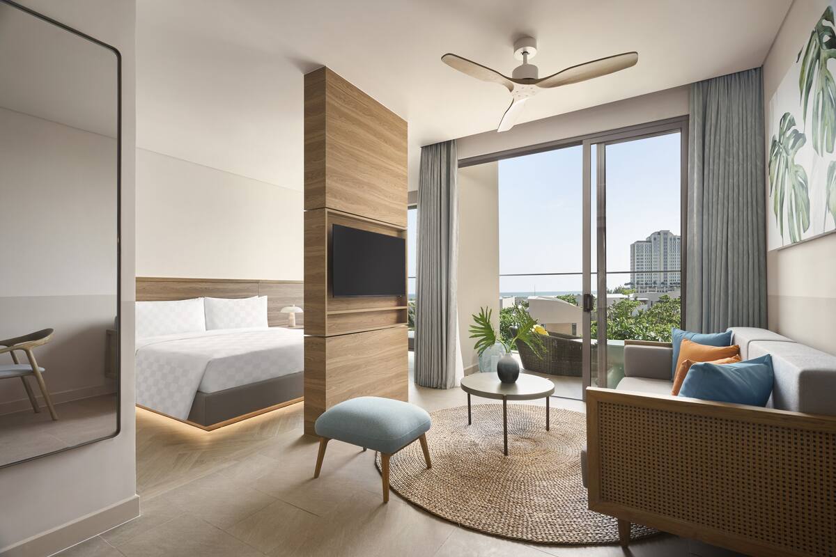 A modern hotel room with a bed, a sofa, a small table, a ceiling fan, and a large window with a city view.