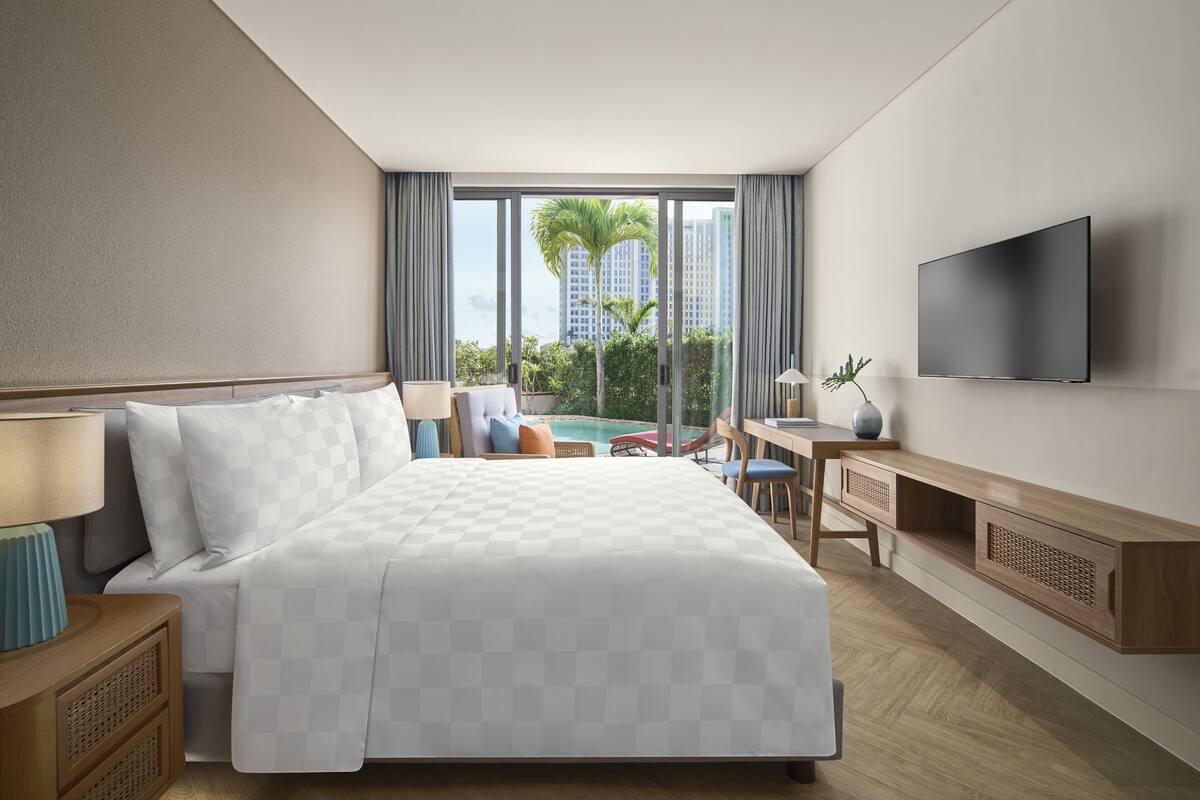 A hotel room with a large bed, a desk, a TV, and a view of palm trees and buildings.