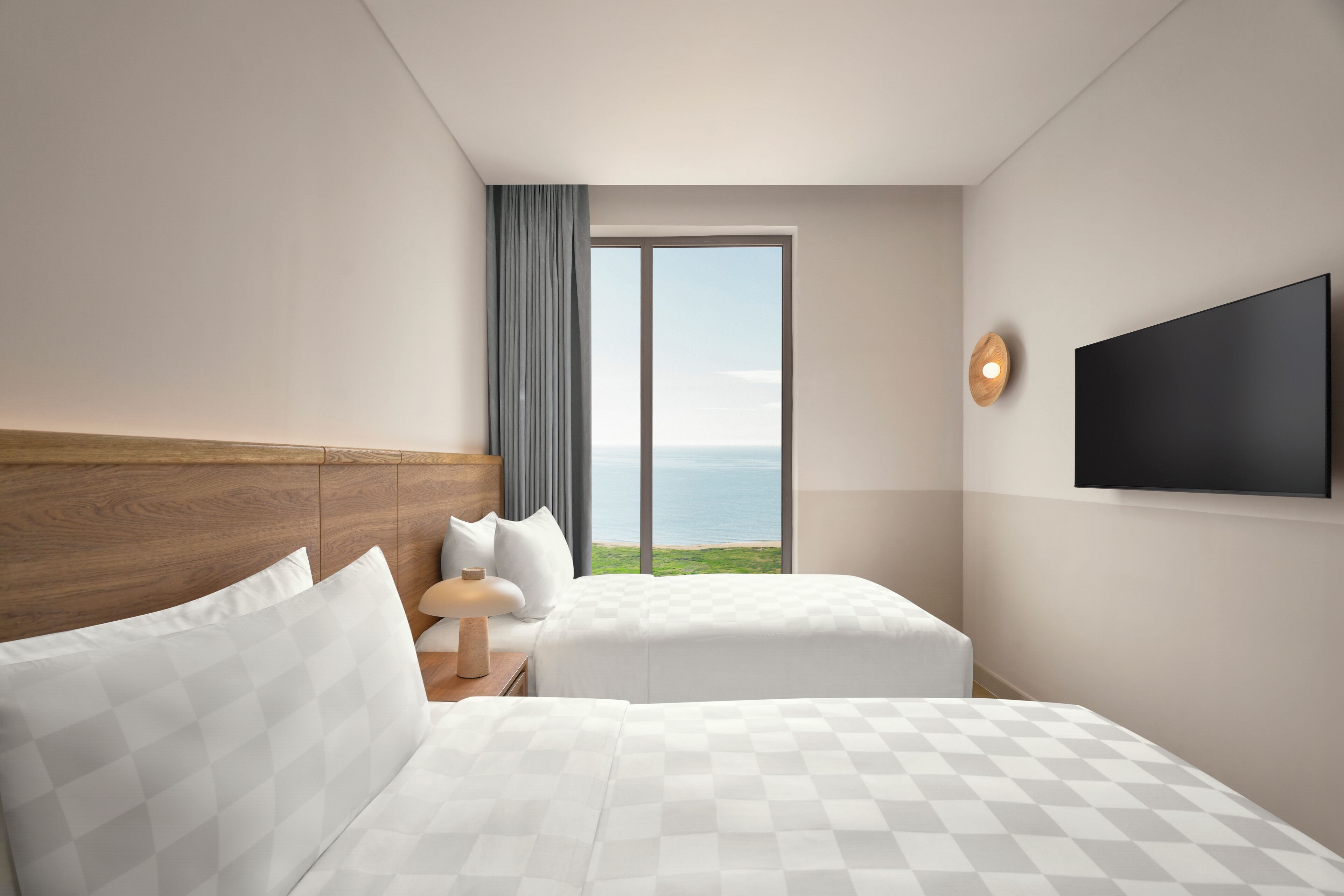 A hotel room with two beds, a large window with a view of the sea, a flat-screen TV, and a wooden headboard.
