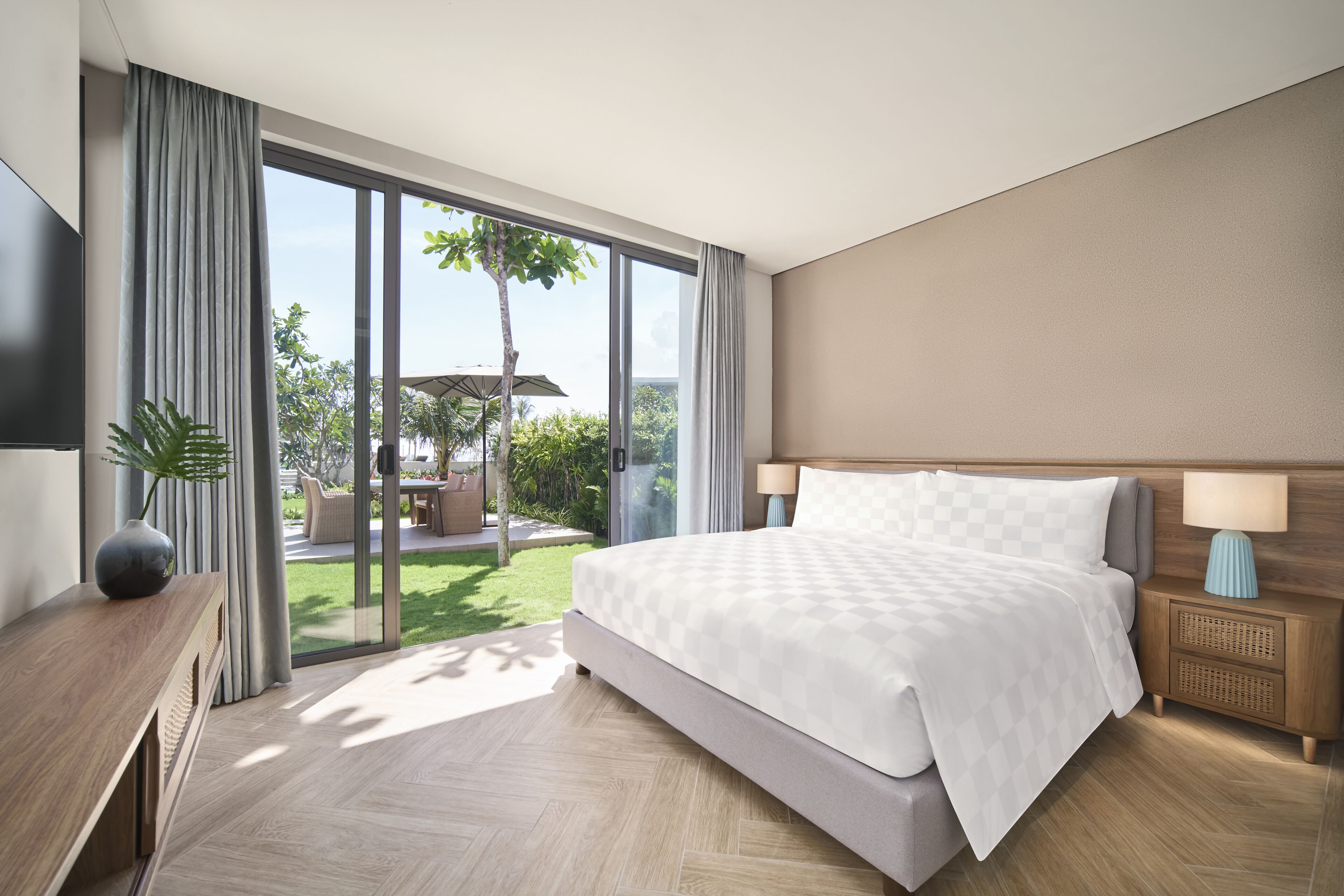Une chambre d’hôtel moderne avec un grand lit, une table de chevet en bois, une télévision et une vue sur un jardin grâce à de grandes fenêtres.