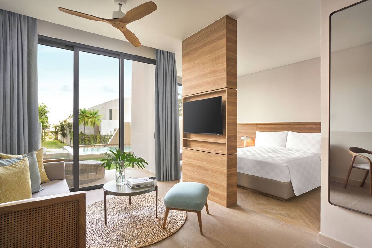A modern hotel room with a large window, a flat-screen TV, a bed, a sofa, a small table, and a ceiling fan.