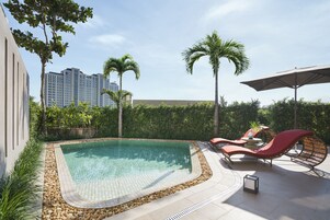 Ein Swimmingpool mit Liegestühlen und Sonnenschirm, umgeben von viel Grün und Palmen.