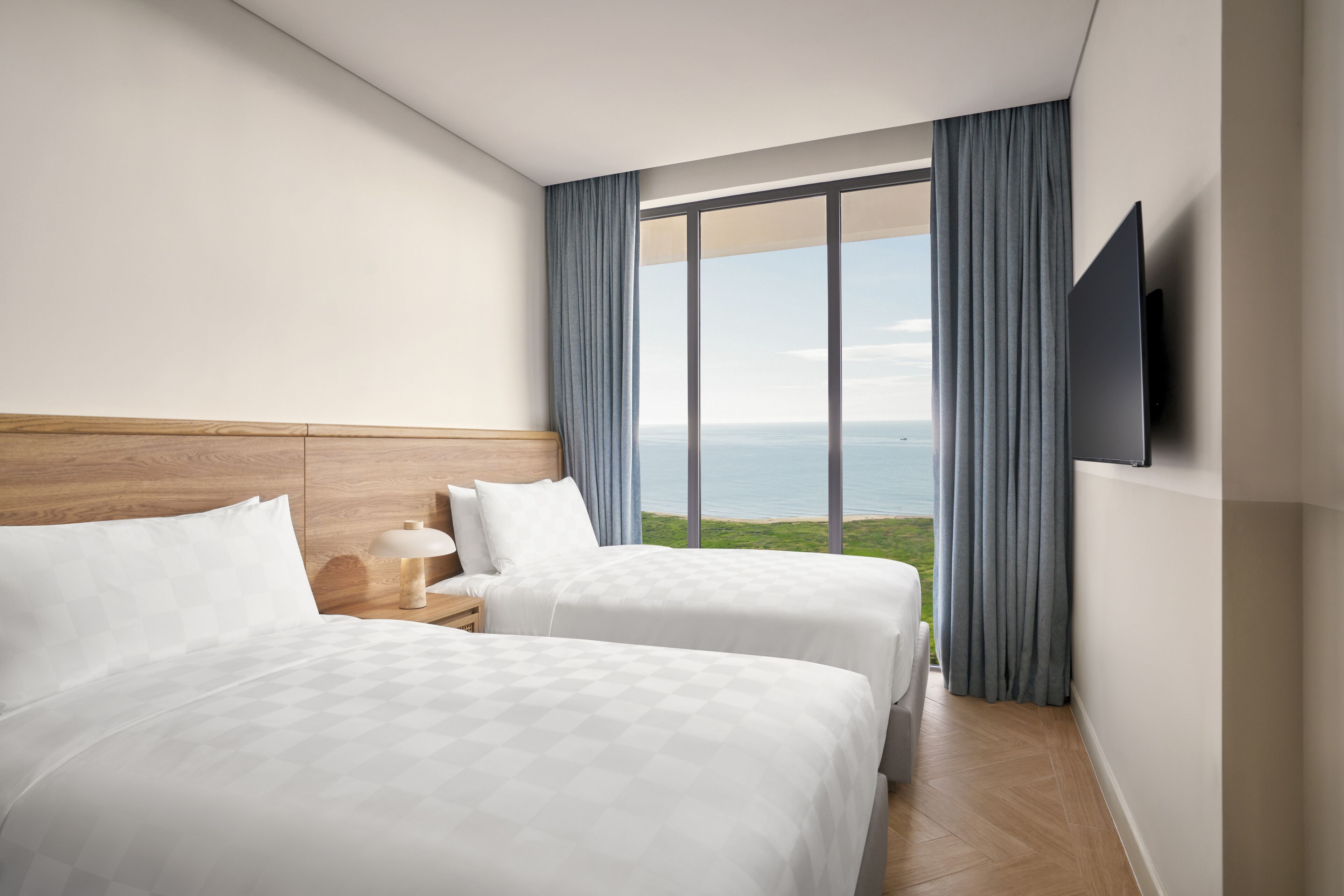 A hotel room with two beds, a wooden headboard, a nightstand with a lamp, a TV mounted on the wall, and a large window with a view of the sea.