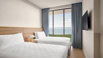 A hotel room with two beds, a wooden headboard, a nightstand with a lamp, a TV mounted on the wall, and a large window with a view of the sea.