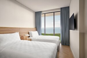 Habitación de hotel con dos camas, cabecera de madera, mesita de noche con lámpara, televisor montado en la pared y un ventanal con vista al mar.