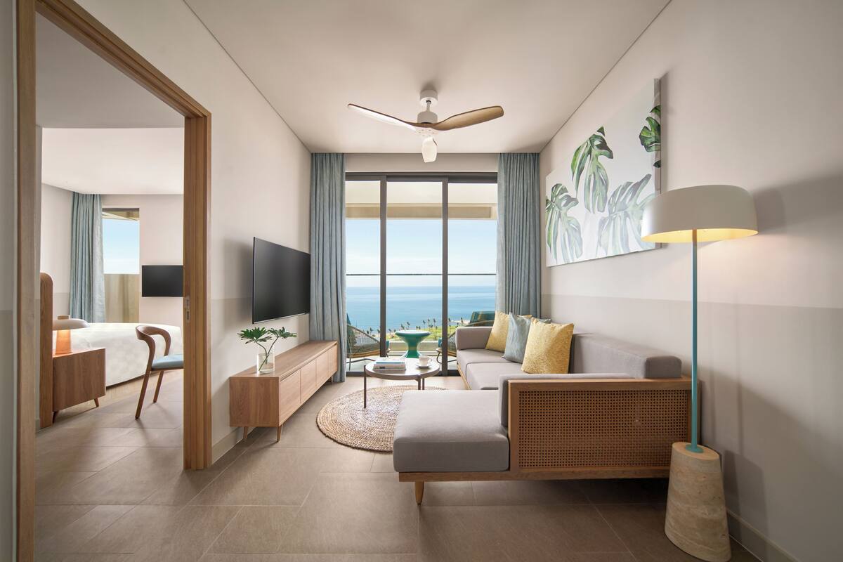 A modern hotel room with a large window, a ceiling fan, a flat-screen TV, a sofa, a coffee table, and a side table with a plant.