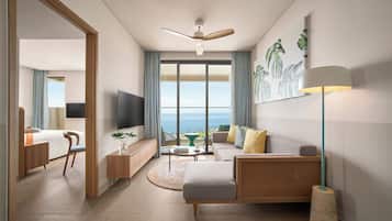 A modern hotel room with a large window, a ceiling fan, a flat-screen TV, a sofa, a coffee table, and a side table with a plant.