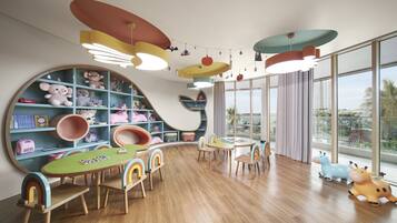 Una sala de juegos infantil con una estantería grande, una mesa de madera con sillas y vistas a palmeras a través de grandes ventanales.
