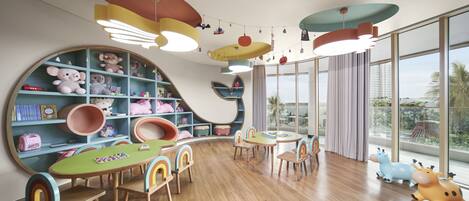 Una sala de juegos infantil con una estantería grande, una mesa de madera con sillas y vistas a palmeras a través de grandes ventanales.
