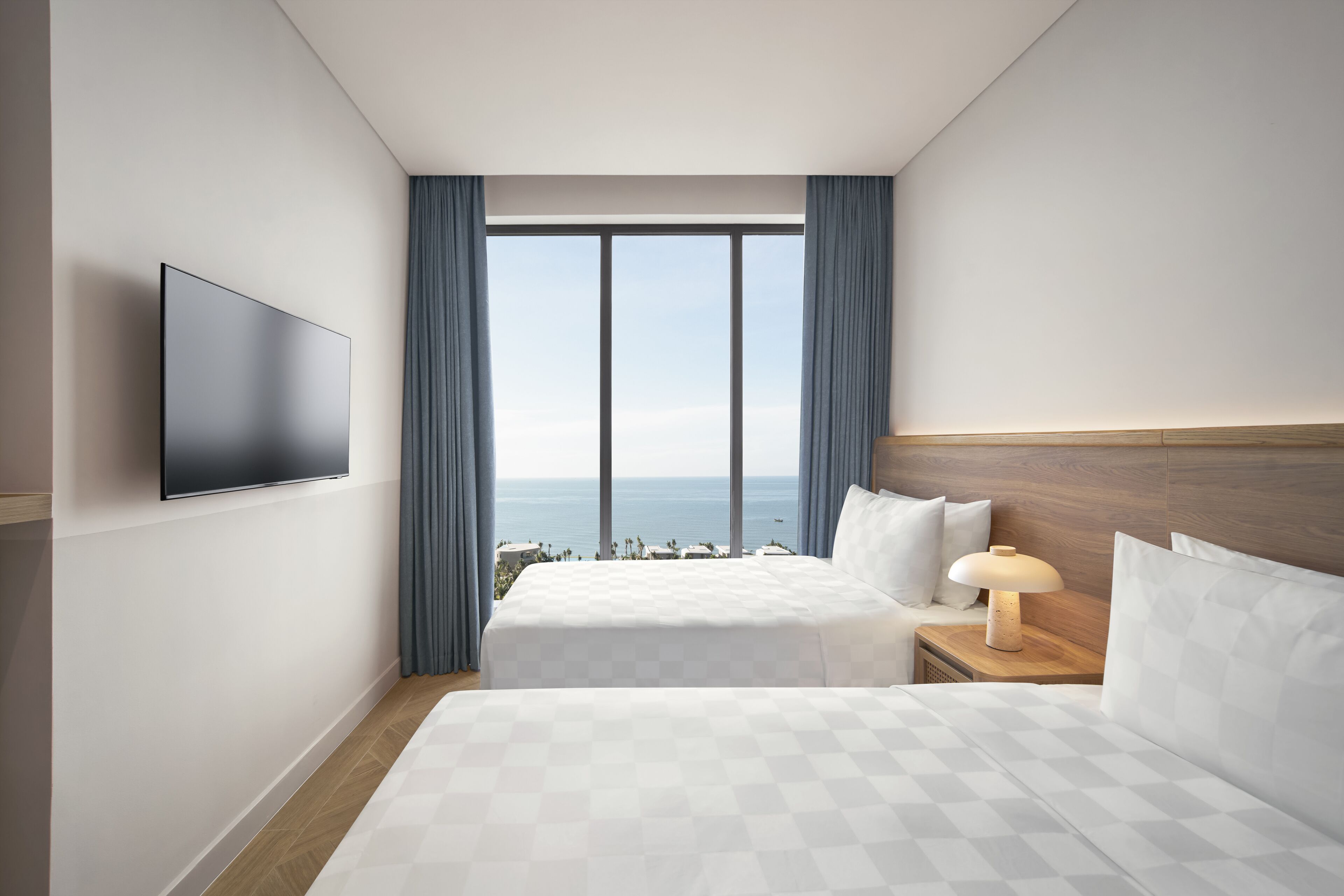 A hotel room with a large bed, a TV mounted on the wall, a nightstand with a lamp, and a view of the ocean through large windows.
