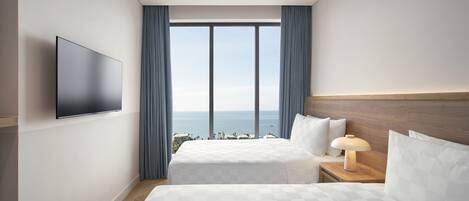 A hotel room with a large bed, a TV mounted on the wall, a nightstand with a lamp, and a view of the ocean through large windows.