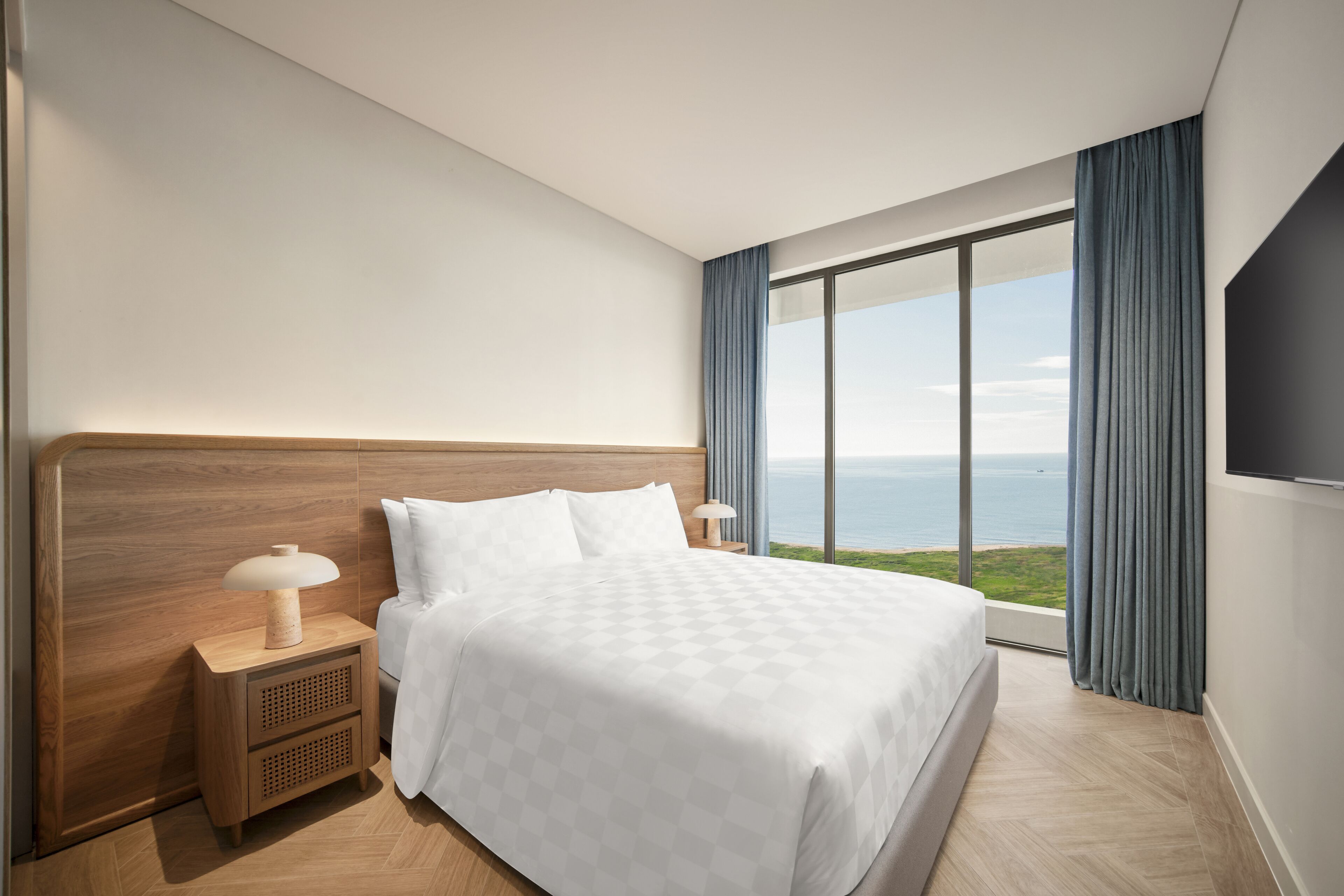 Habitación de hotel con una cama grande, una mesita de noche con lámpara, un televisor en la pared y vistas al mar.