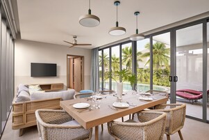 Un comedor moderno con una mesa de madera dispuesta para cuatro, rodeada de sillas de mimbre, un televisor de pantalla plana en la pared y vistas a una piscina y palmeras a través de amplias puertas de cristal.