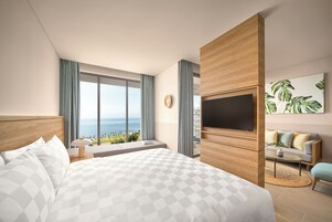 A modern hotel room with a large bed, a TV, a sofa, and a view of the ocean.