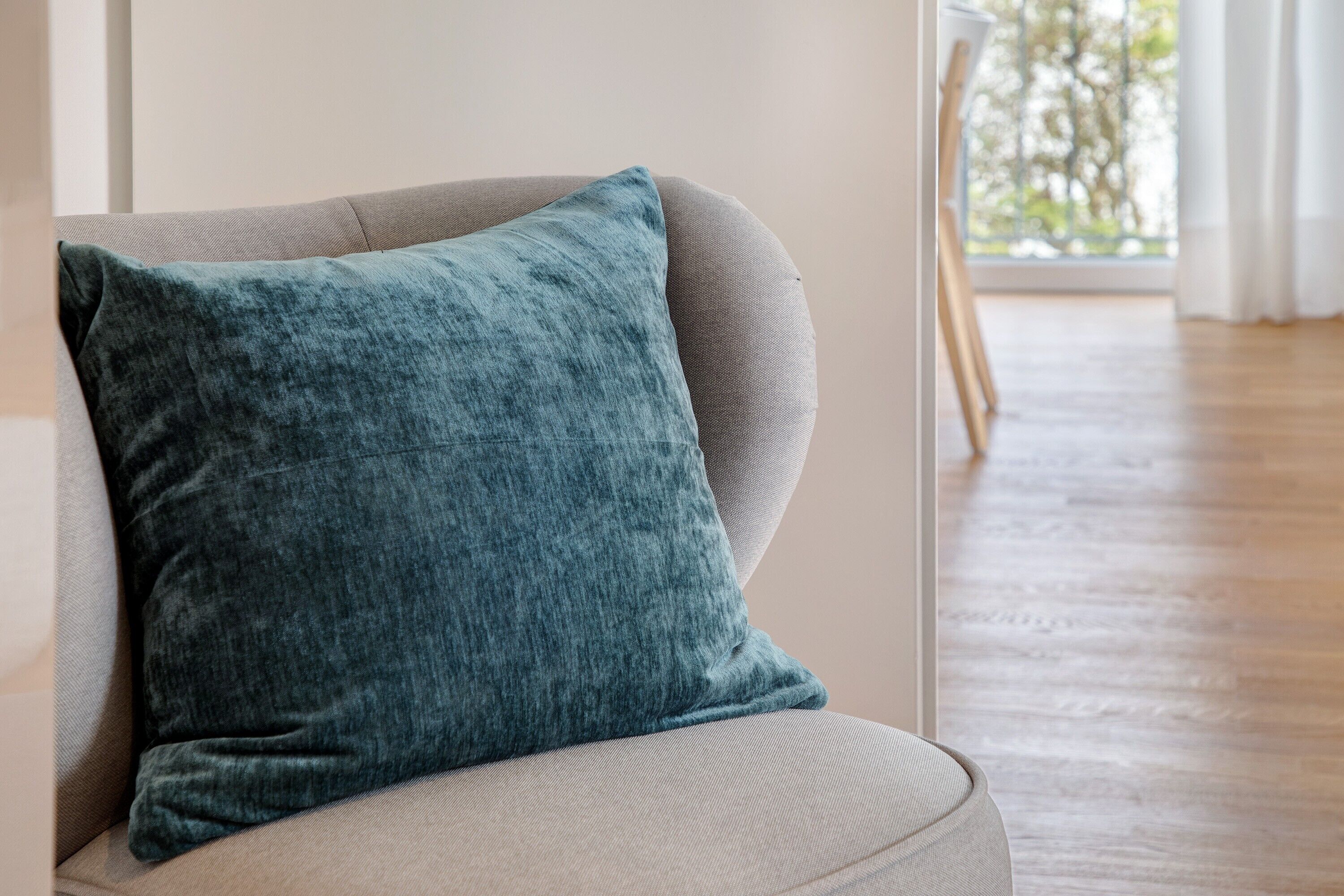 Une chaise avec un coussin de couleur turquoise, dans une pièce dotée d’un plancher en bois et d’une fenêtre donnant sur un paysage verdoyant.