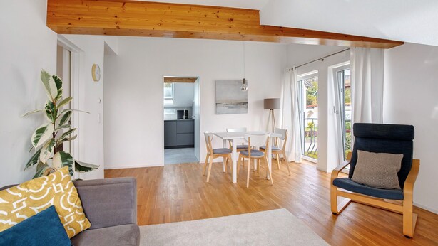 Een moderne woonkamer met een houten plafond, een grijze bank, een blauwe fauteuil, een eettafel met stoelen en een potplant.