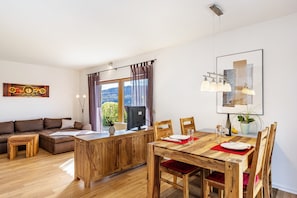 A modern living room with a wooden dining table, a sofa, and a large window with curtains.