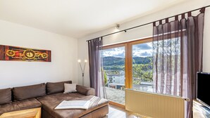 A modern living room with a brown sectional sofa, a wooden coffee table, a large window with curtains, a radiator, and a flat-screen TV. - Apartment "Südalb" with Shared Garden & Wi-Fi (Albstadt)