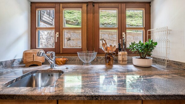 A kitchen with granite countertops, a stainless steel sink, wooden cabinets, a stone backsplash, and a view through large French doors.