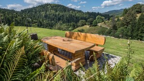 A wooden picnic table with benches in a grassy field with a forested hill in the background.