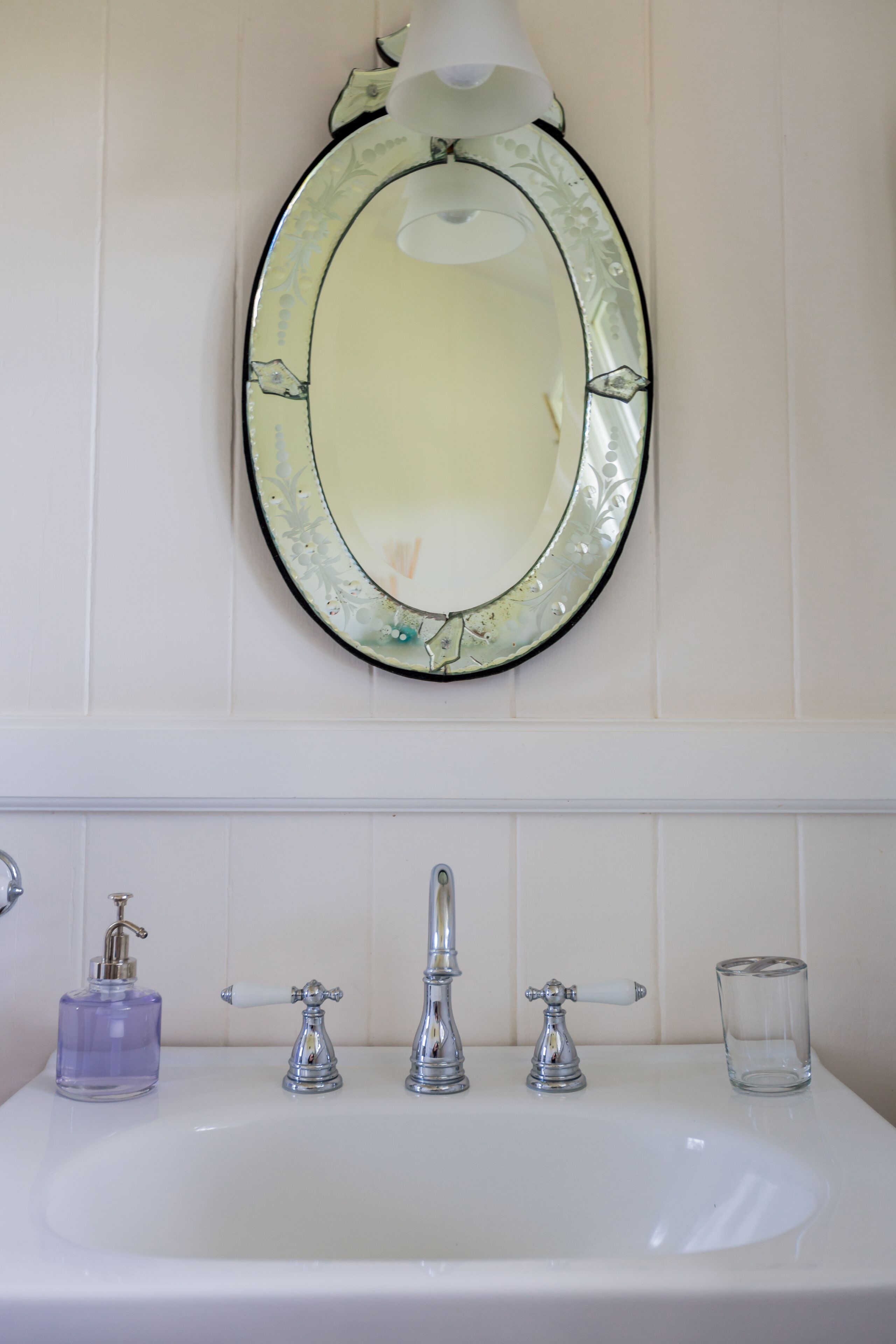 A bathroom with a white sink, three faucets, a purple soap dispenser, and a clear glass.