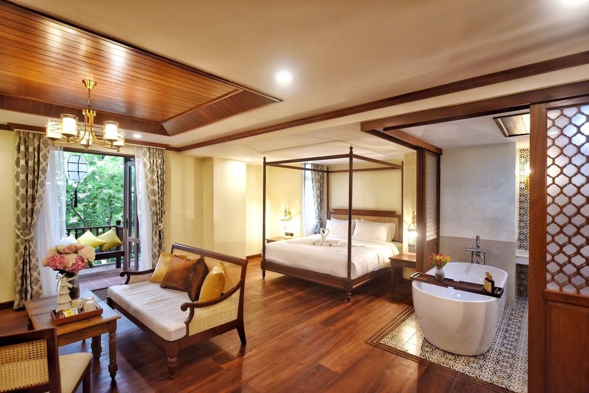 Un lit à baldaquin, un coin salon avec un canapé et des fauteuils, une grande fenêtre avec des rideaux et une salle de bain séparée avec une baignoire.