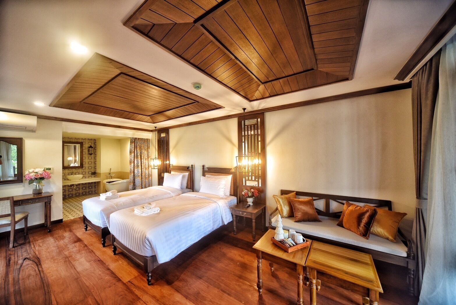 Habitación de hotel con dos camas, una zona de estar, tocador con espejo y techo de madera.